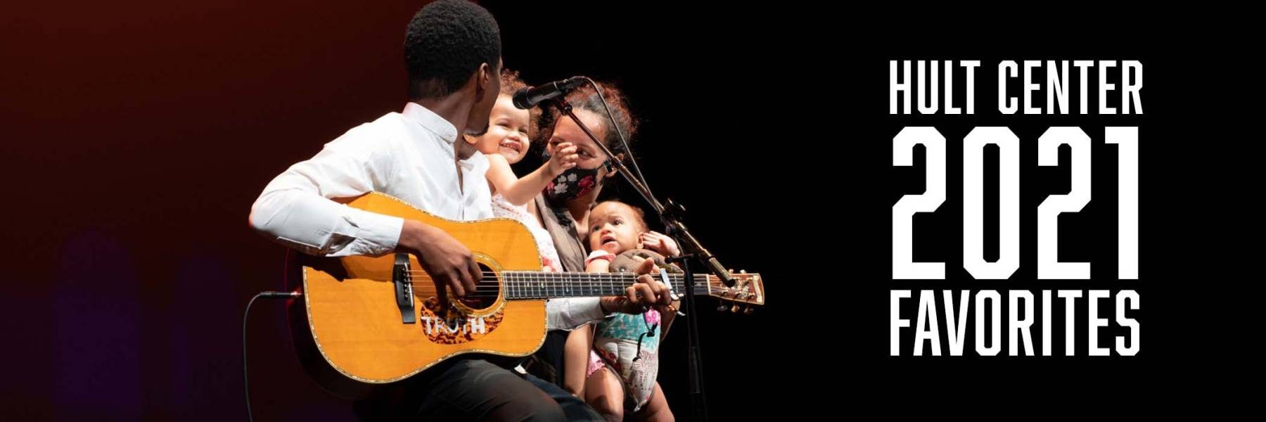 A man sitting on a stool on stage holding a guitar and a microphone in front of him with his wife and two children beside him.