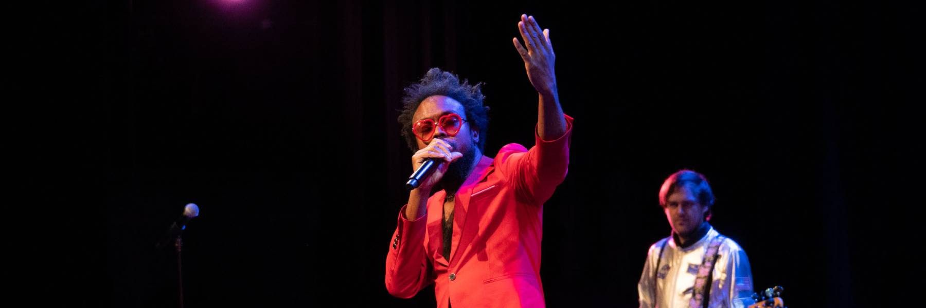A man in a bright colored suit and heart shaped sunglasses singing into a microphone and a musician in the background.
